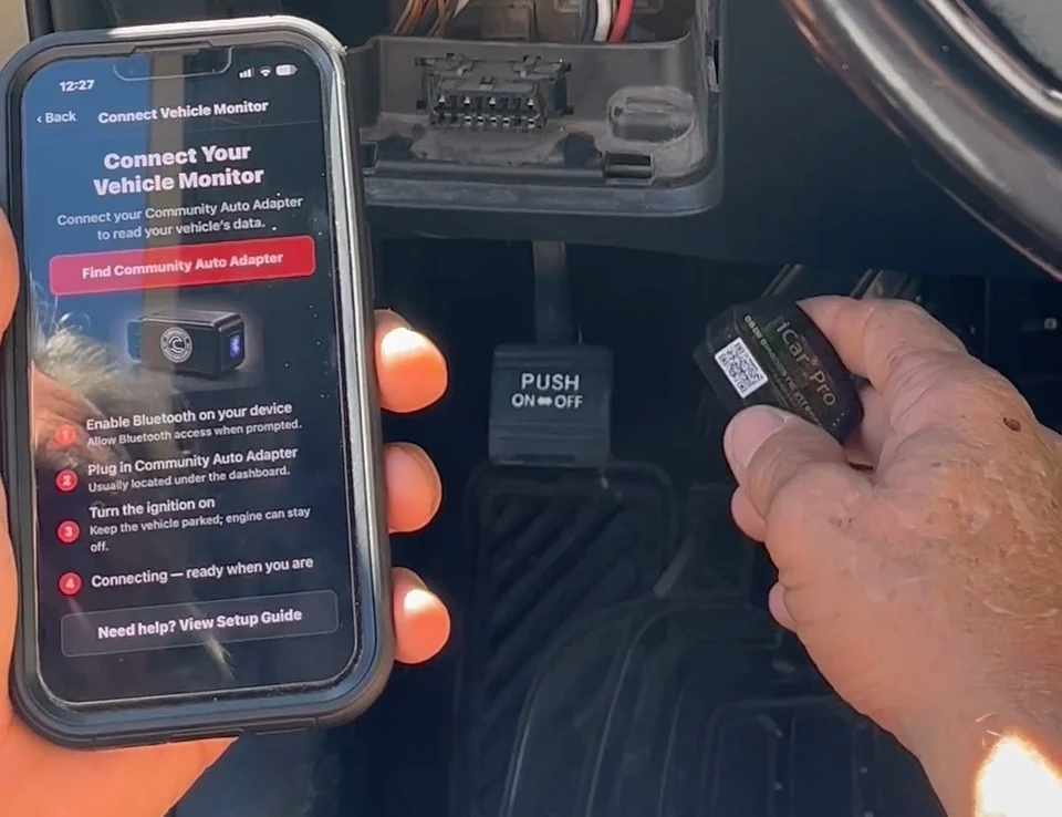 A physical phone running Community Auto beside a Bluetooth vehicle adapter inside a vehicle